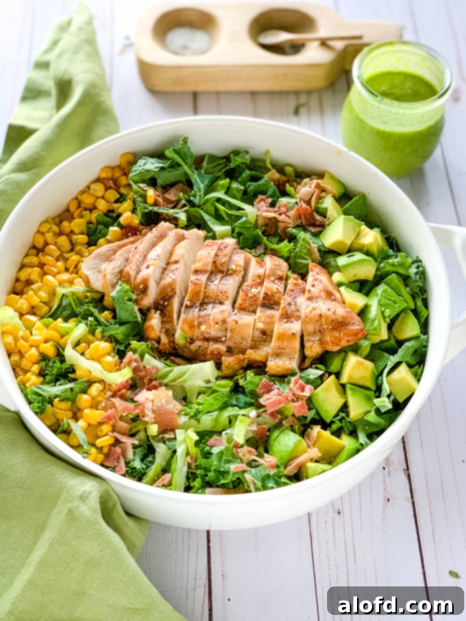 Sun-Kissed Suppers Made Simple 9 A refreshing chicken cobb salad served in a white bowl, elegantly placed next to a green linen napkin, salt and pepper cellars, and a Weck jar filled with salad dressing.