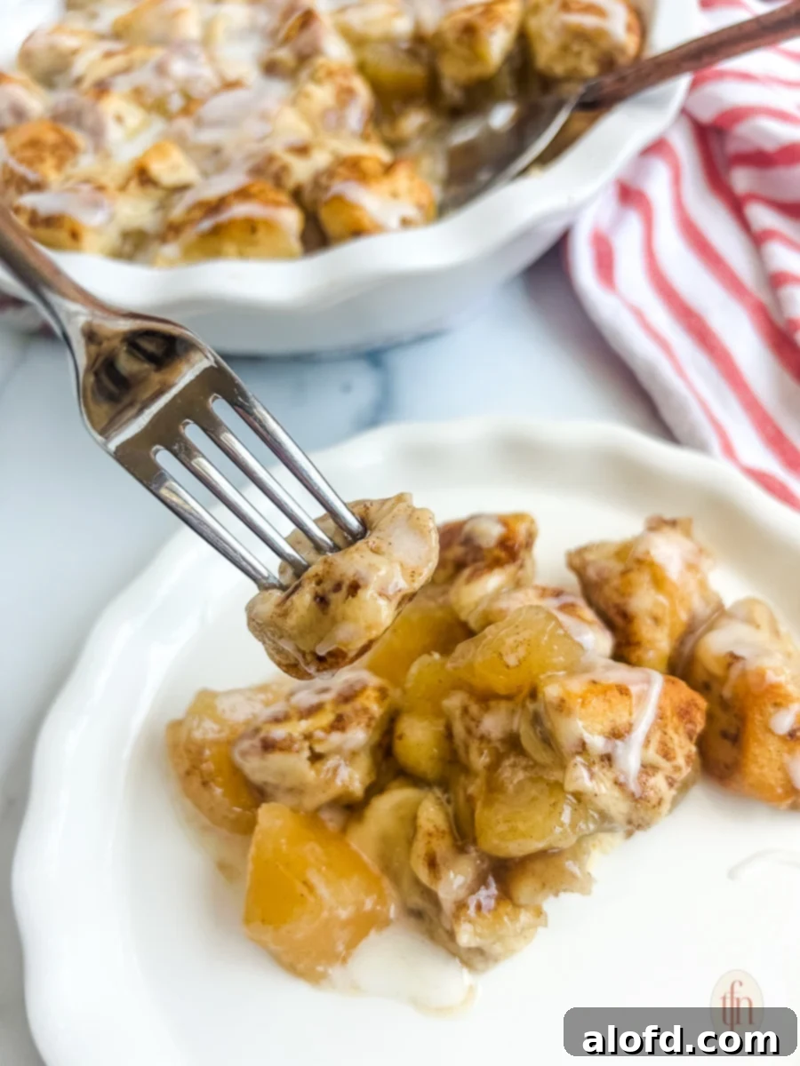 Pieces of apple pie filling and cinnamon roll on a fork held above a white plate, ready to be enjoyed.