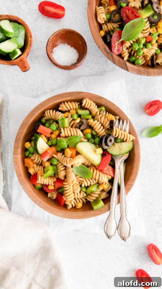 Lunchbox Hacks for Educators 14 A vibrant rainbow pasta salad and silverware elegantly arranged on a wooden bowl, with another bowl of salad, salt, cucumber slices, and cherry tomatoes artfully placed on a white cloth in the background.