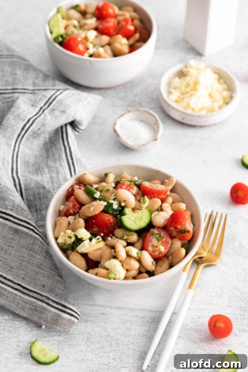 Lunchbox Hacks for Educators 22 A pristine white bowl filled with cannellini bean salad, flanked by forks. Chopped tomatoes, cucumber, another bowl of salad, a gray cloth, and a bowl of feta are artfully arranged in the background, completing a fresh and inviting scene.