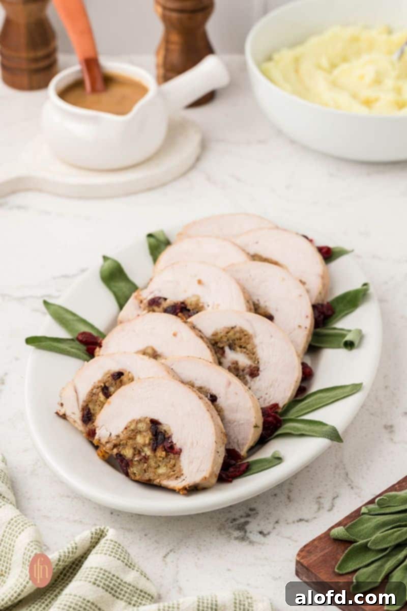 A wide, eye-level shot of a long white platter with seven slices of stuffed turkey breast, garnished with sage and dried cranberries. A gravy boat and a bowl of mashed potatoes are in the soft-focus background.
