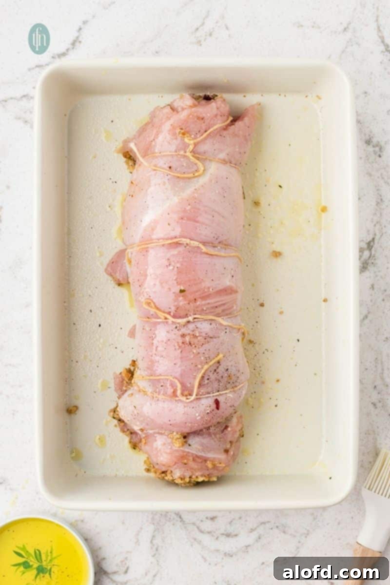 An overhead shot of the raw stuffed turkey breast, rolled up and tied with kitchen twine, resting in a white rectangular baking dish, lightly coated with oil.