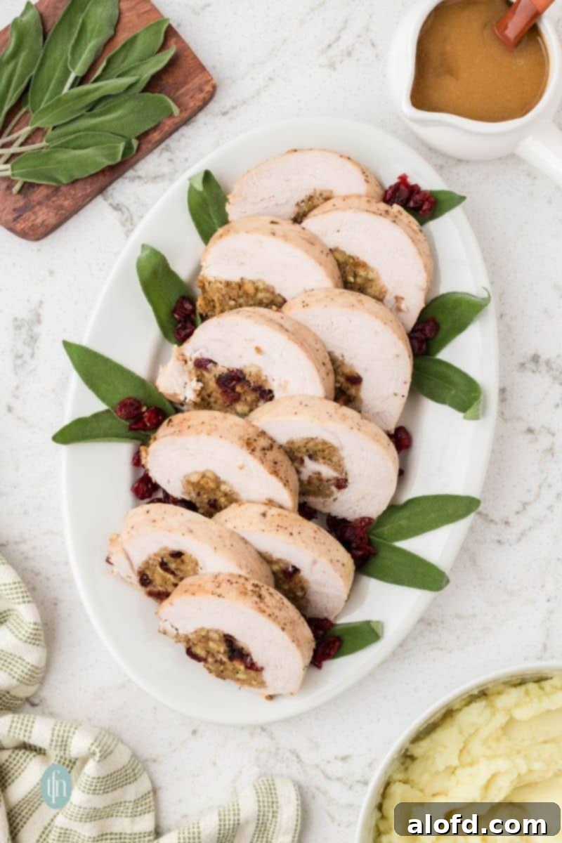 An overhead shot of a long white platter holding seven slices of baked, stuffed turkey breast garnished with fresh sage leaves and dried cranberries. A gravy boat and bowl of mashed potatoes are visible on the counter.