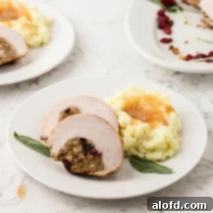 A square-cropped image of two slices of stuffed turkey breast served on a white plate next to a mound of mashed potatoes topped with gravy and garnished with a sage leaf.
