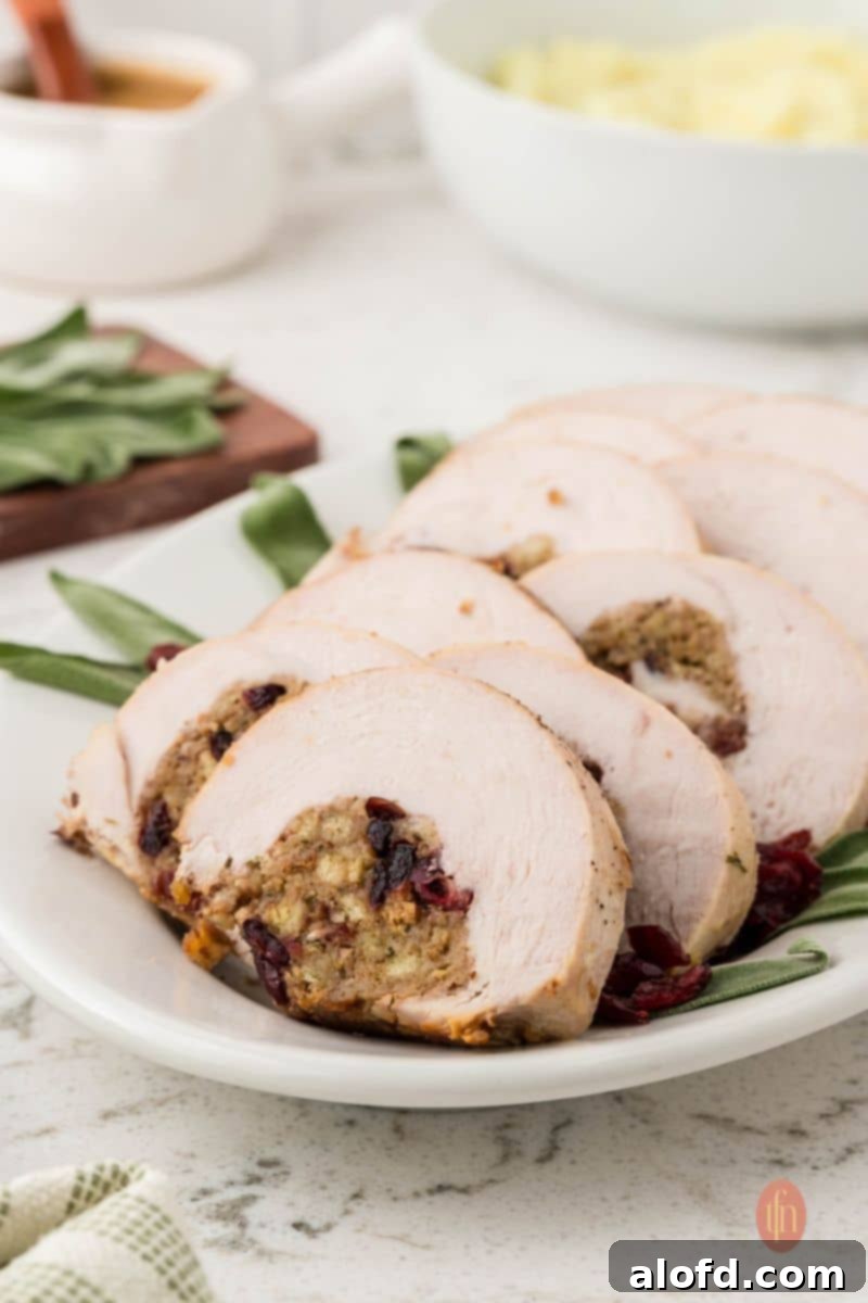 A slightly angled, eye-level shot of a long white platter with seven slices of stuffed turkey breast garnished with sage leaves and dried cranberries. A gravy boat and bowl of mashed potatoes are in the background.