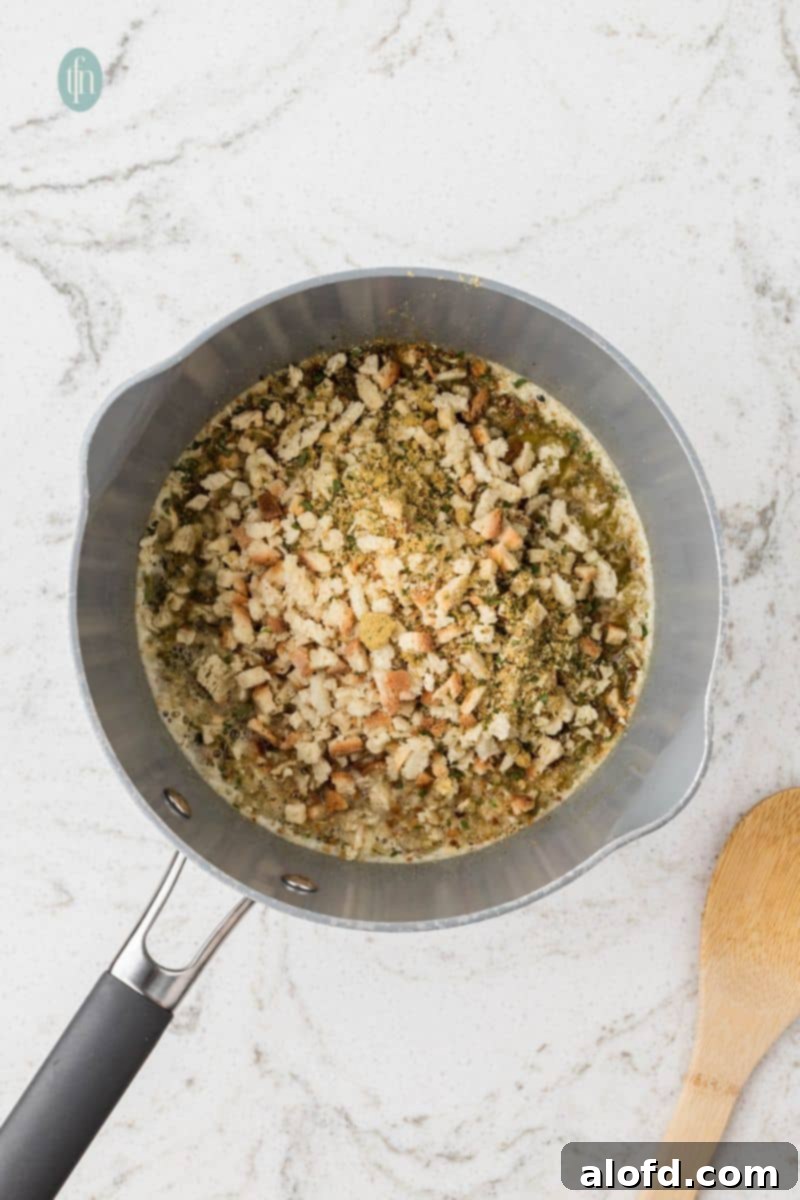 An overhead shot of a saucepan where small bread cubes and dried herbs have been added to a seasoned liquid, beginning to form the stuffing mixture.
