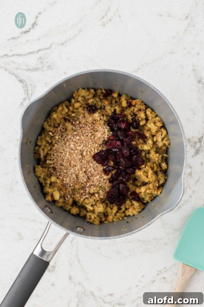 An overhead shot of a saucepan filled with the stuffing mixture, topped with dried cranberries and crushed pecans or walnuts, ready to be mixed.