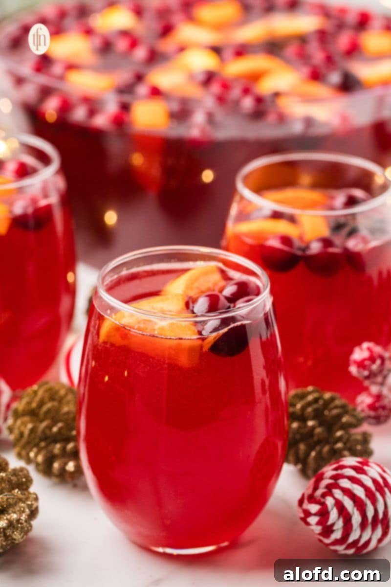 Close-up of glasses filled with vibrant red Christmas punch, garnished with orange slices and cranberries. A large punch bowl is blurred in the background, set with festive gold and red ornaments.