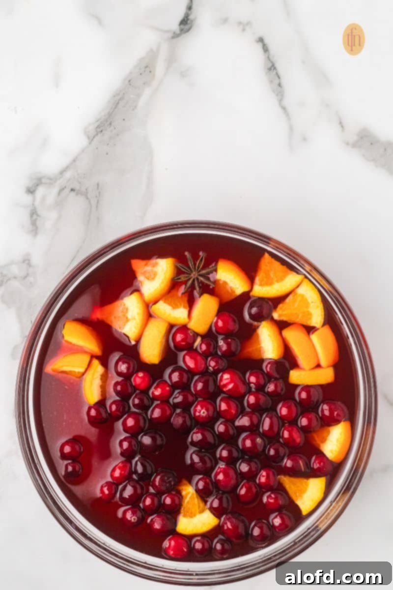 Overhead view of a punch bowl filled with red punch, currently being garnished with cranberries, orange slices, and star anise before serving.