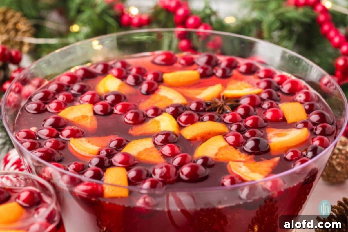 Close-up of a festive Christmas punch in a glass bowl, showing the deep red liquid with floating whole cranberries, orange segments, and star anise against a blurred holiday backdrop.