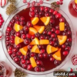 Overhead view of a large glass bowl of holiday punch garnished with floating cranberries, orange slices, and a star of anise. The bowl is surrounded by Christmas decor like gold pinecones and red bead garlands.