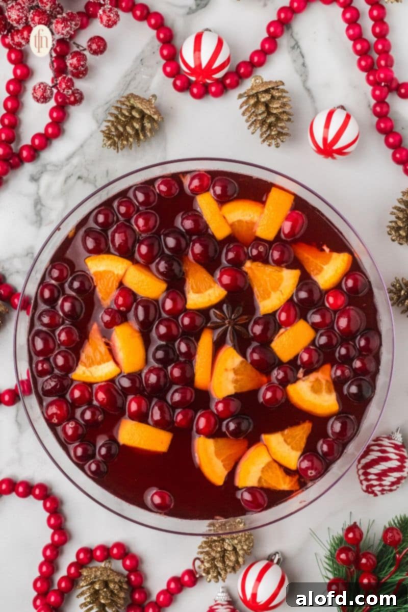Overhead view of a large glass bowl of holiday punch garnished with floating cranberries, orange slices, and a star of anise. The bowl is surrounded by Christmas decor like gold pinecones and red bead garlands.