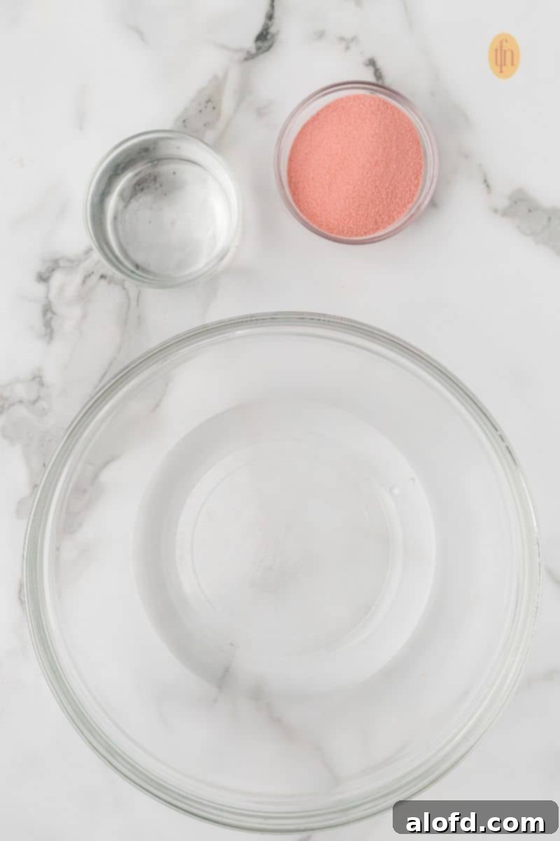 Overhead view of the initial punch ingredients: an empty mixing bowl, a small bowl of water, and a small bowl of cherry gelatin powder on a white marble surface.