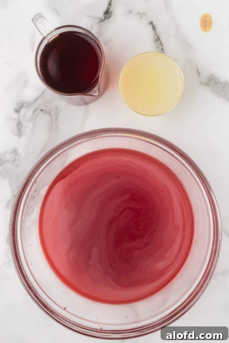 Overhead shot showing a large glass bowl with a swirling red gelatin mixture, a glass measuring cup of dark red juice, and a small bowl of lemonade concentrate on a white marble surface.