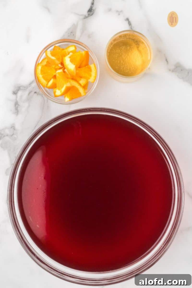 Overhead view of ingredients for punch: a large glass bowl containing the red juice mixture, a bowl of orange slices, and a small glass of ginger ale or sparkling cider on a white marble background.