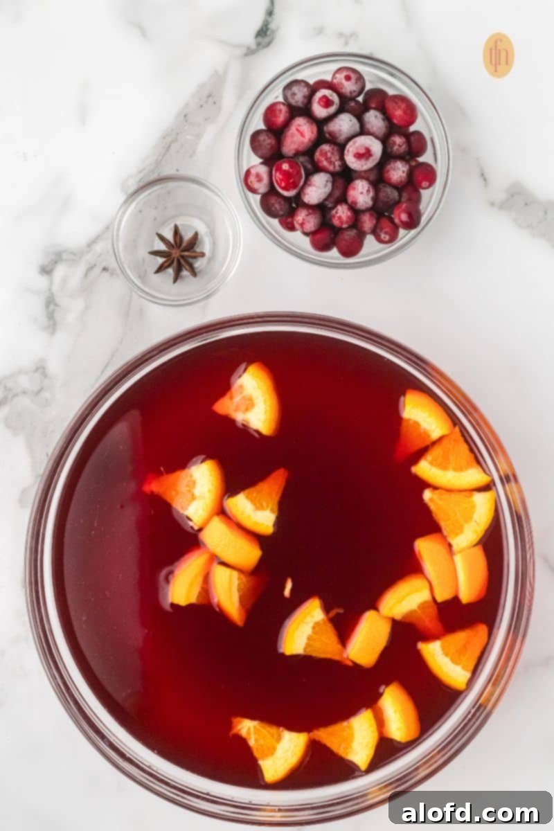 Ingredients for punch: a glass bowl of red liquid with orange slices, a bowl of frozen cranberries, and a small bowl with a star of anise on a marble counter.