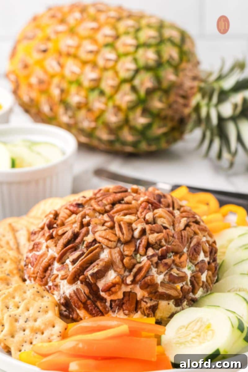 The pecan-crusted cheese ball appetizer served on a platter with sliced cucumbers and orange bell peppers in the foreground, with a whole fresh pineapple sitting on the counter in the background. The ultimate party appetizer.