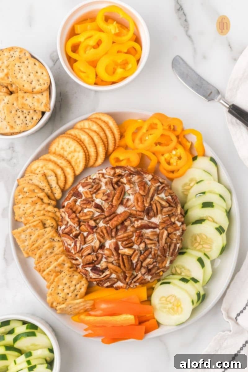 Overhead view of a completed pecan cheese ball appetizer on a platter, surrounded by circular rows of round crackers, sliced cucumbers, and yellow and orange bell pepper rings.