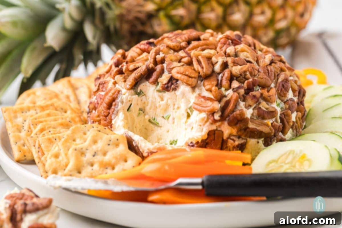 A close-up shot of a pecan-crusted cheese ball with a section scooped out, showing the creamy interior mixed with herbs or pineapple, served with crackers and vegetables.