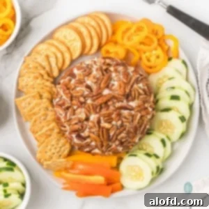 A round cream cheese ball coated entirely in pecans, served on a white platter with crackers, sliced cucumbers, and rings of yellow and orange bell peppers.