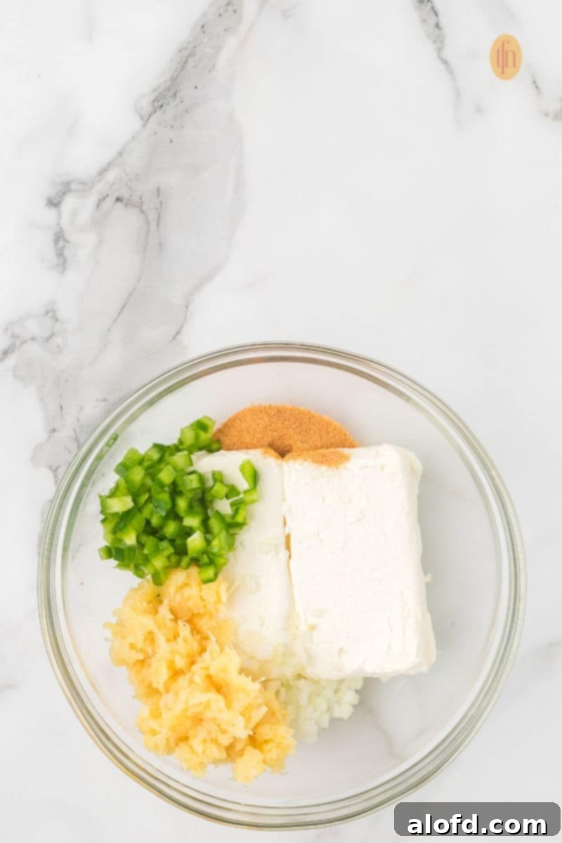 A clear glass bowl containing the raw ingredients for the cheese ball: a block of cream cheese, chopped green peppers, crushed pineapple, and spices/seasonings.