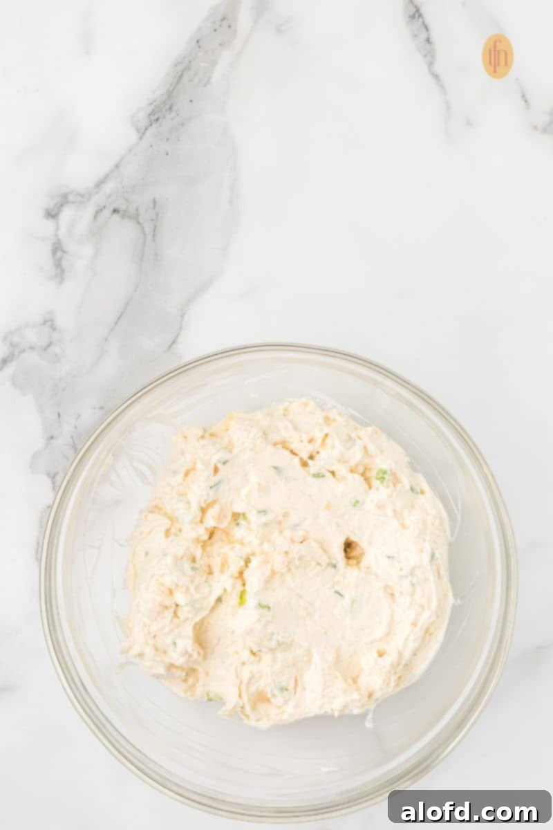 A clear glass bowl holding the fully mixed cheese ball ingredients—a creamy, off-white mixture with visible green pieces of seasoning or pepper.