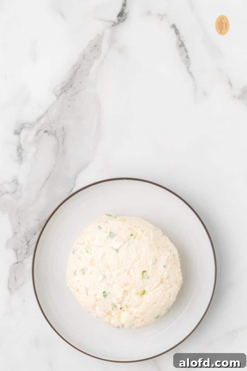 Top-down view of a round, smooth cream cheese ball (without a pecan coating) resting on a small white plate with a brown rim, set against a marble background.
