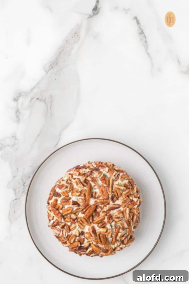 Top-down view of a cheese ball that has just been coated entirely with pecan halves, resting on a small white plate against a marble countertop.