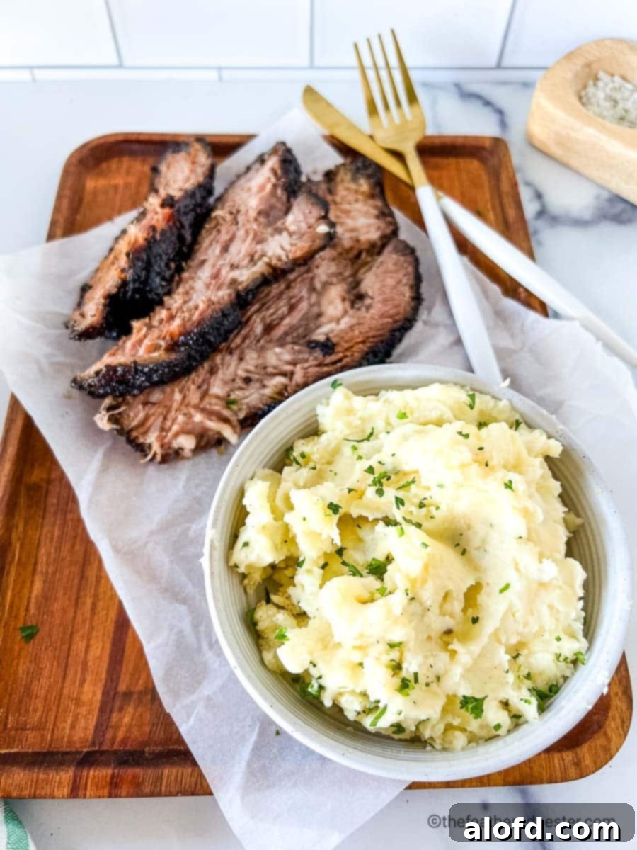 Dreamy Instant Pot Red Potato Mash 2 A white bowl of red skin mashed potatoes next to slices of brisket on a wooden serving board with a striped napkin and gold fork and knife in the background.