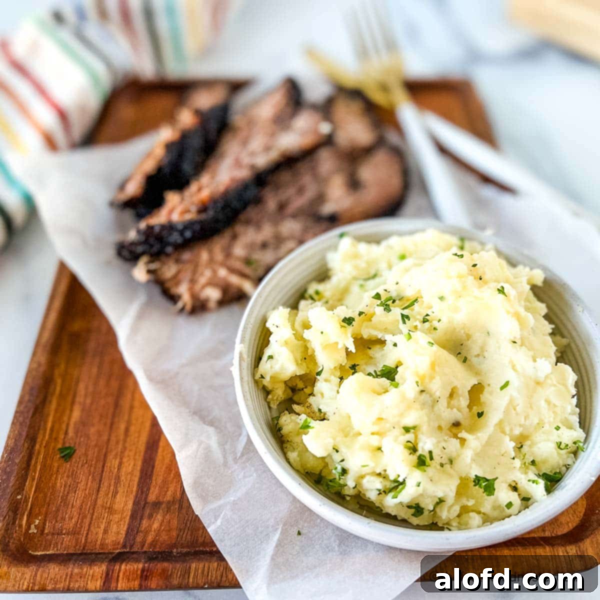 Dreamy Instant Pot Red Potato Mash 9 a white bowl of red mashed potatoes with slices of brisket on a wooden serving board with a striped napkin and gold fork and knife in the background.