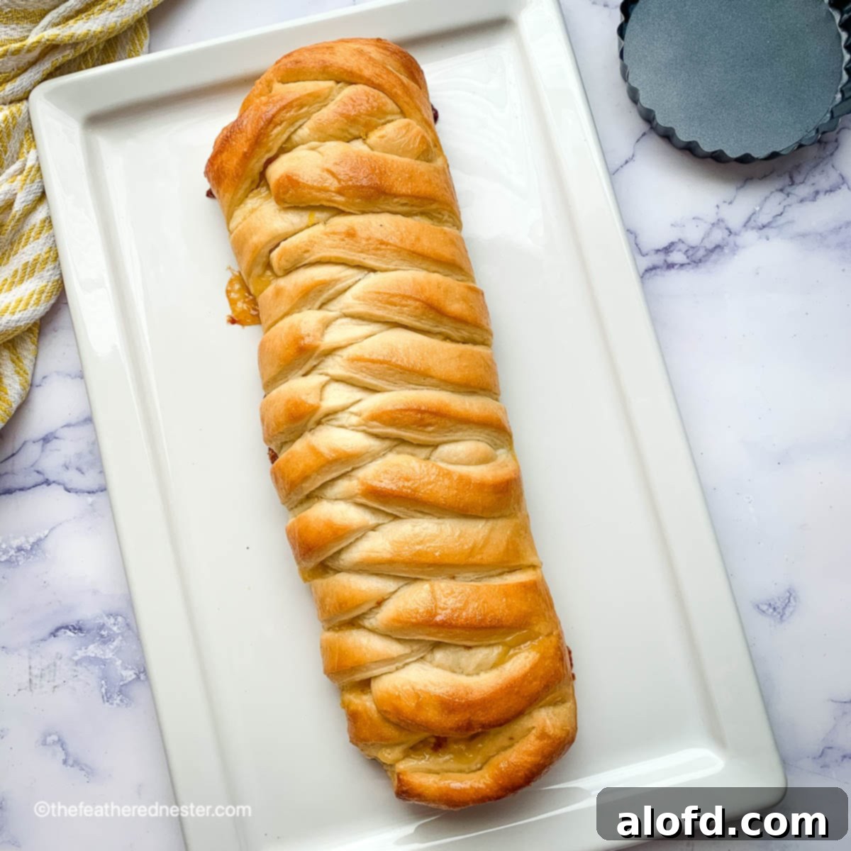 Lemon curd and cream cheese braided bread loaf on a white platter with yellow and white towel and mini tart pan, showcasing its golden crust and delicious filling.