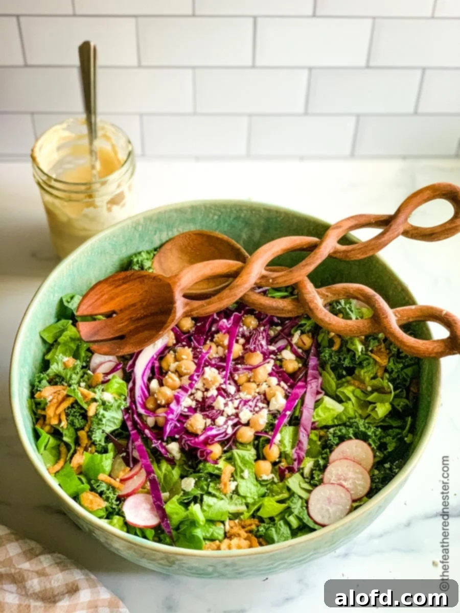 Healthy fresh greens in a green bowl with radishes, chickpeas, feta, and wood tongs. A jar of dressing sits behind the bowl.