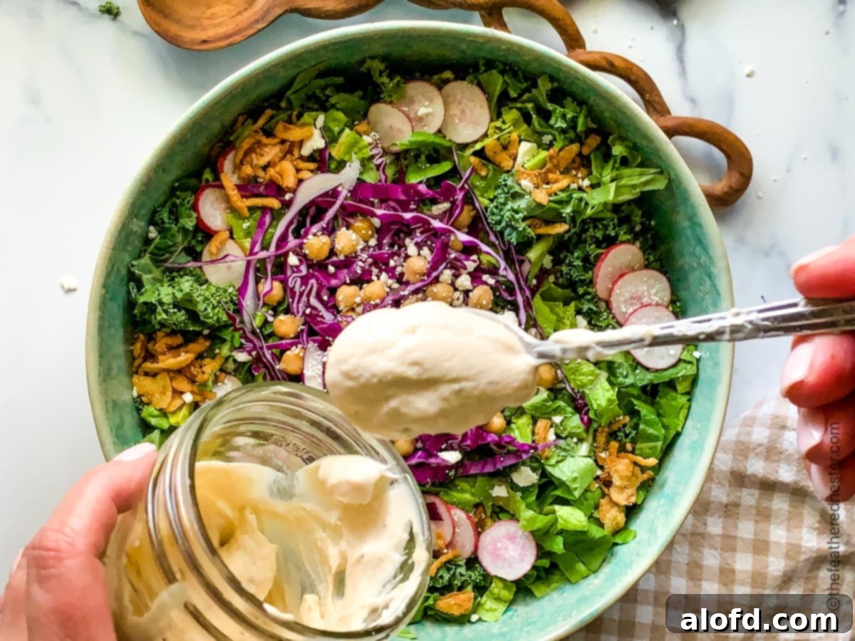 A large serving bowl filled with vibrant Greek Kale Salad, featuring fresh romaine, tender kale, thinly sliced radishes, and creamy feta cheese, being drizzled generously with homemade tahini dressing. Salad tongs rest beside the bowl.