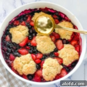 a white casserole dish with strawberry and blueberry cobbler and a gold serving spoon.
