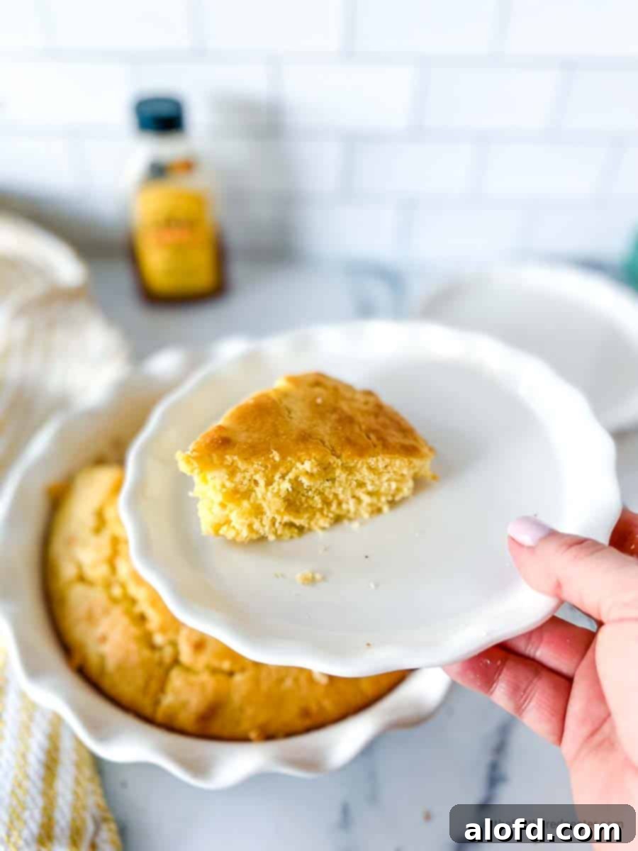 a hand holding a white plate with a slice of Jiffy cornbread with a casserole dish of cornbread, yellow striped cloth, plate, and a bottle of honey in the background.