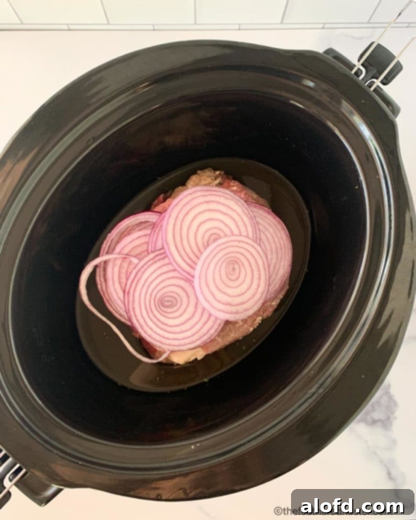 Meltingly Tender Slow Cooker Chuck Roast 5 A slow cooker with a ranch-seasoned chuck roast submerged in beef broth, topped with sliced onions, ready for cooking.