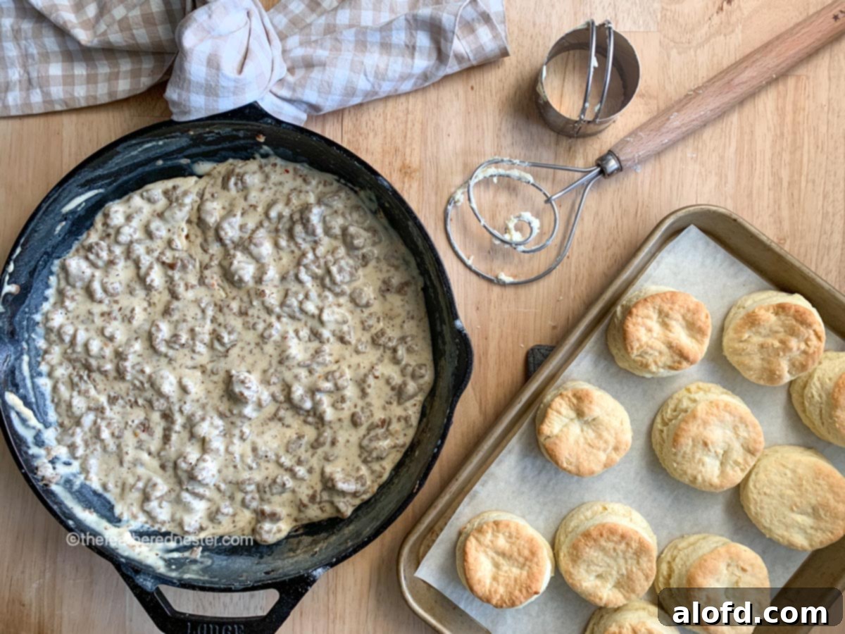 Fluffy Biscuits with Creamy Sausage Gravy 7 a cast iron skillet of sausage gravy and a baking sheet of homemade biscuits with a dough whisk, biscuit cutter, and checked napkin.
