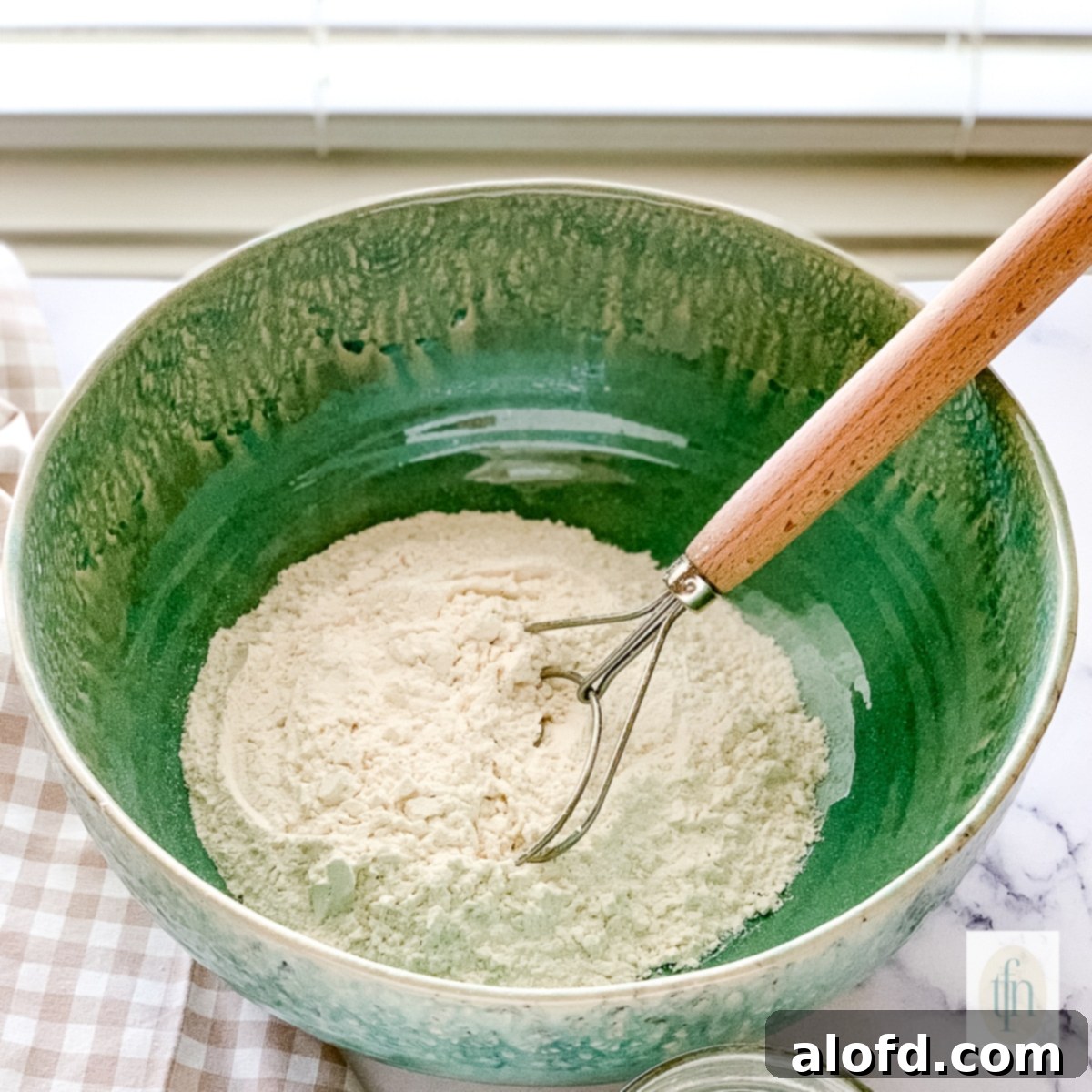 A green bowl filled with dry ingredients, being stirred with a dough whisk.