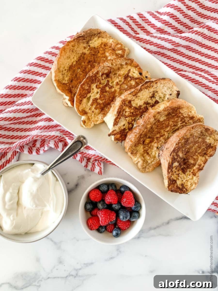 Tangy Sourdough French Toast 2 A white platter with slices of sourdough bread French toast, a small bowl of berries and whipped cream over a red and white napkin.