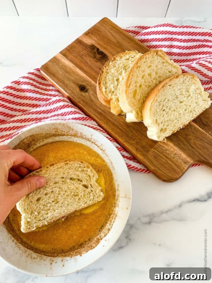 Tangy Sourdough French Toast 5 A slice of sourdough French bread being dipped into an egg batter with a wooden board of sliced bread on top of a red and white napkin in the background.