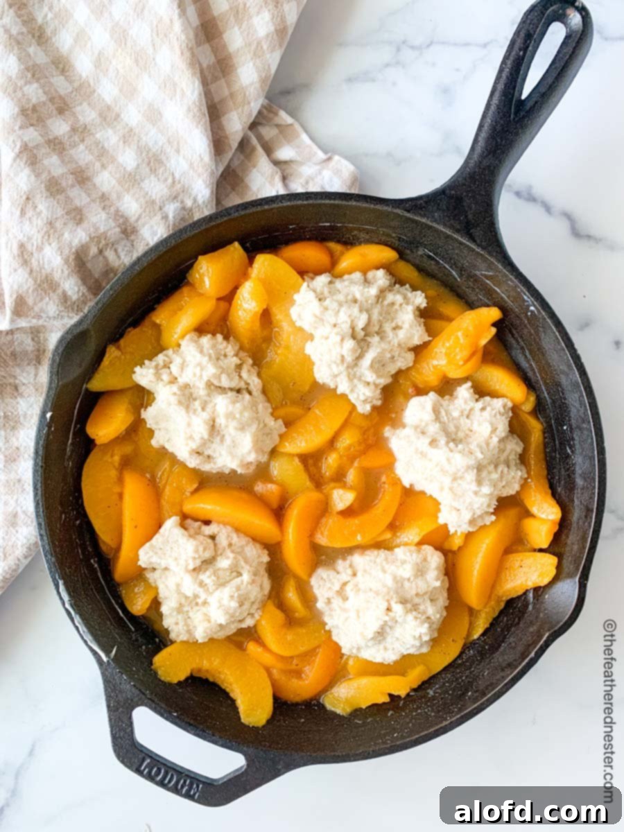 Easy Bisquick Peach Cobbler 6 An unbaked peach cobbler in a cast iron skillet, with dollops of biscuit topping spread over the fruit filling, ready for the oven.