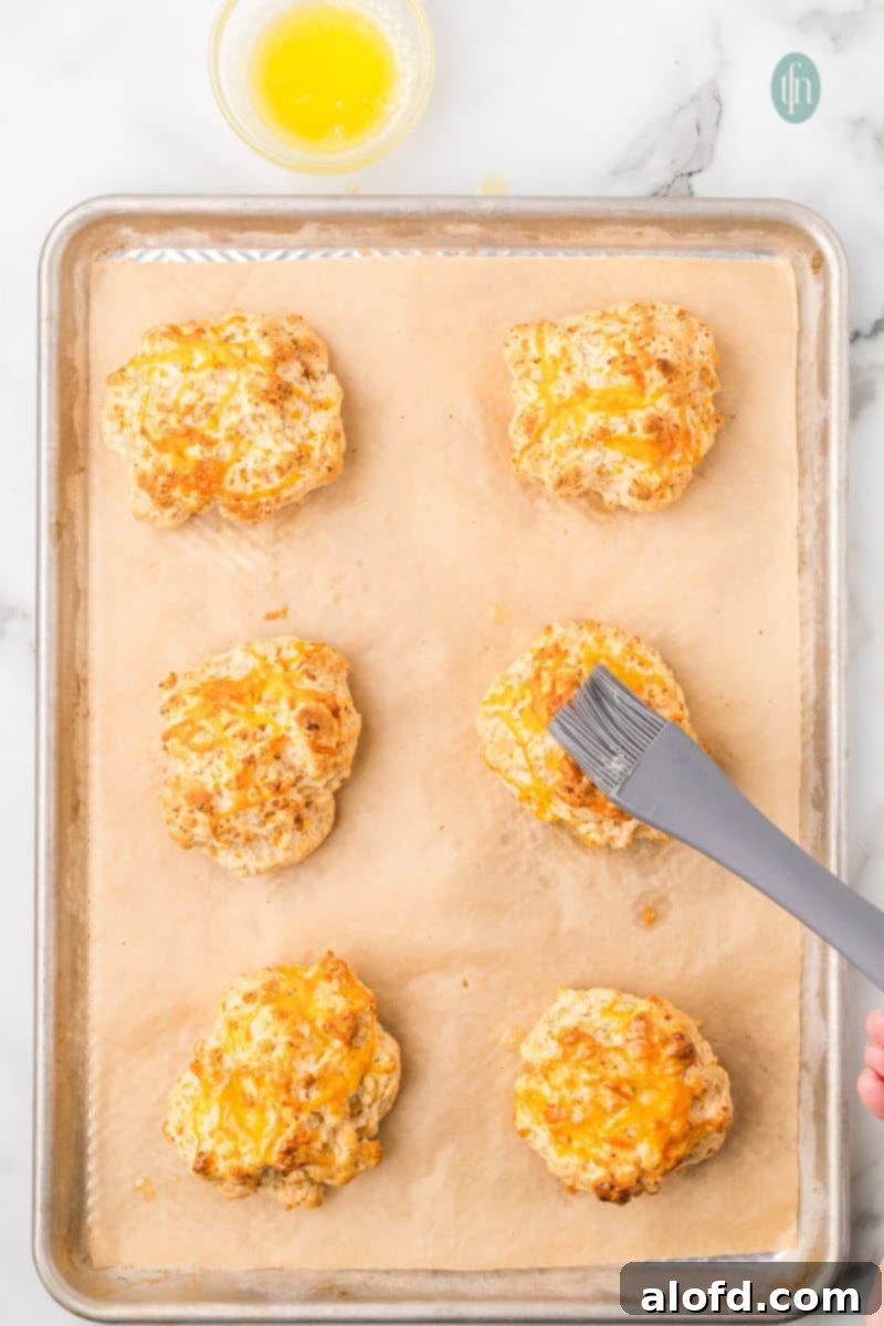 Bisquick Garlic Cheddar Biscuits 14 Freshly baked cheddar biscuits being brushed with melted butter on a baking sheet.