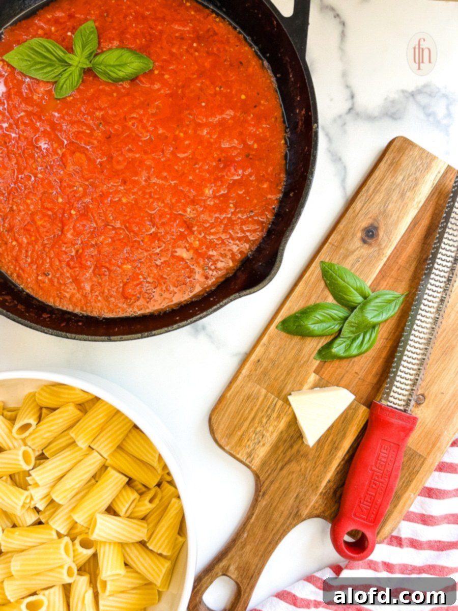 Tomato sauce in a pan, bowl of pasta with sauce over the top, and a wooden cutting board with cheese, grater, and basil sprigs. 