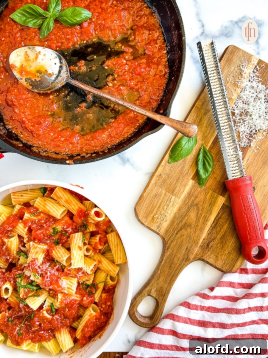 Pasta sauce in a pan, bowl of pasta with sauce over the top, and a wooden cutting board with freshly grated cheese, a grater, and fresh basil leaves.