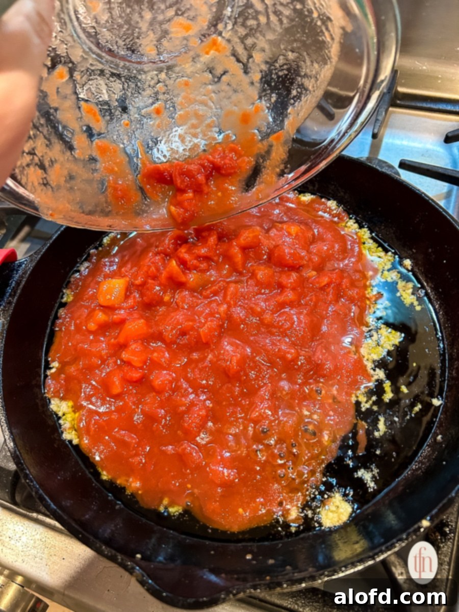 Pouring tomatoes into a pan with toasted garlic on the stove.
