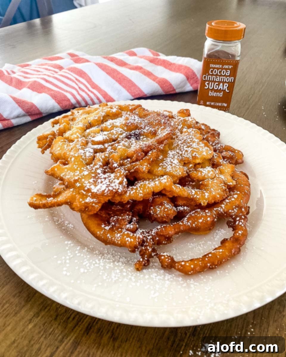 a white plate with a golden-brown Bisquick funnel cake, adorned with powdered sugar, alongside a red striped cloth and a cinnamon powder bottle in the background.