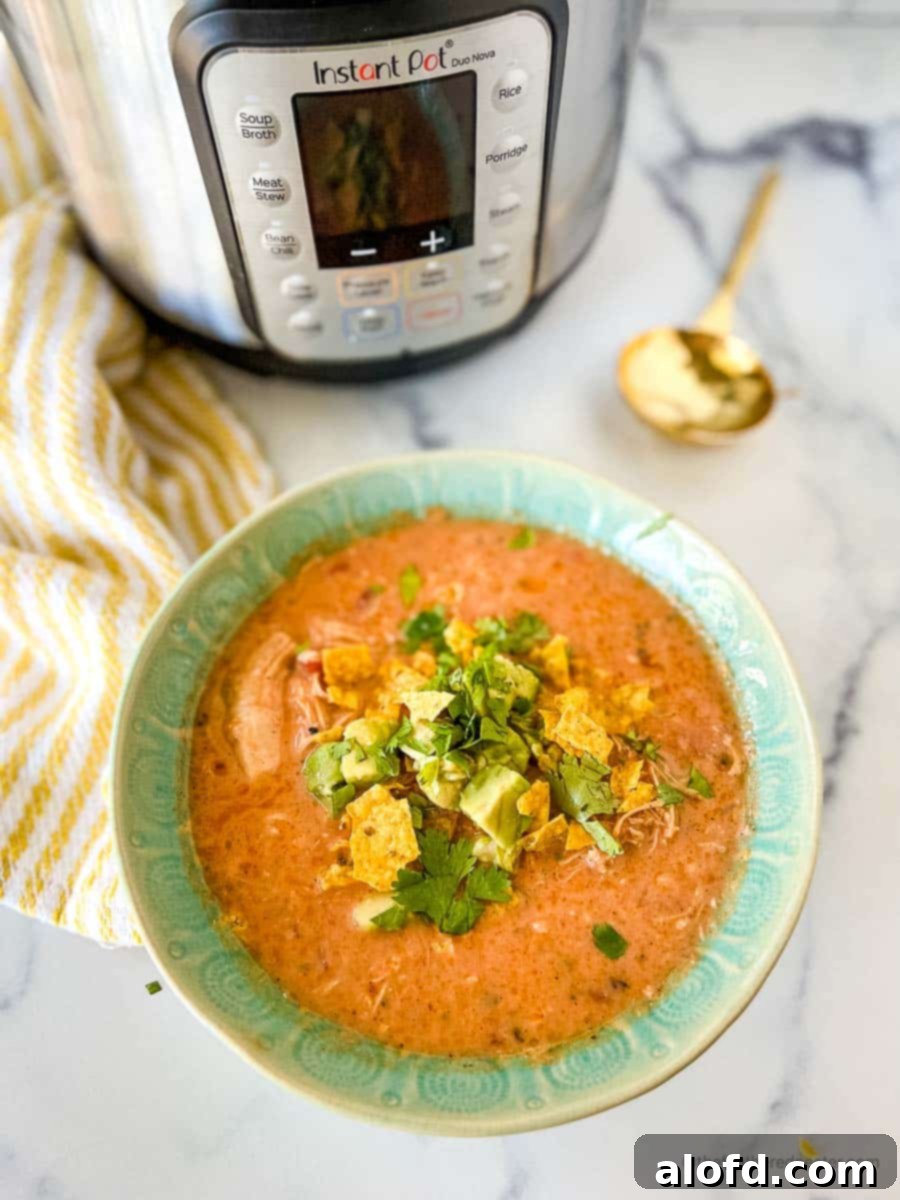 A vibrant green bowl of Instant Pot creamy chicken tortilla soup, garnished with fresh cilantro and crunchy tortilla strips, beside an Instant Pot and a golden spoon on a white and yellow napkin.
