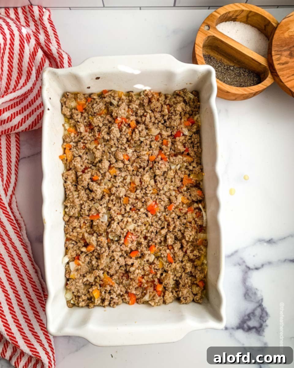 Ultimate Hash Brown Breakfast Bake 7 A casserole dish with a layer of cooked ground sausage, bell pepper, and onion spread over the hash browns, ready for the egg mixture.