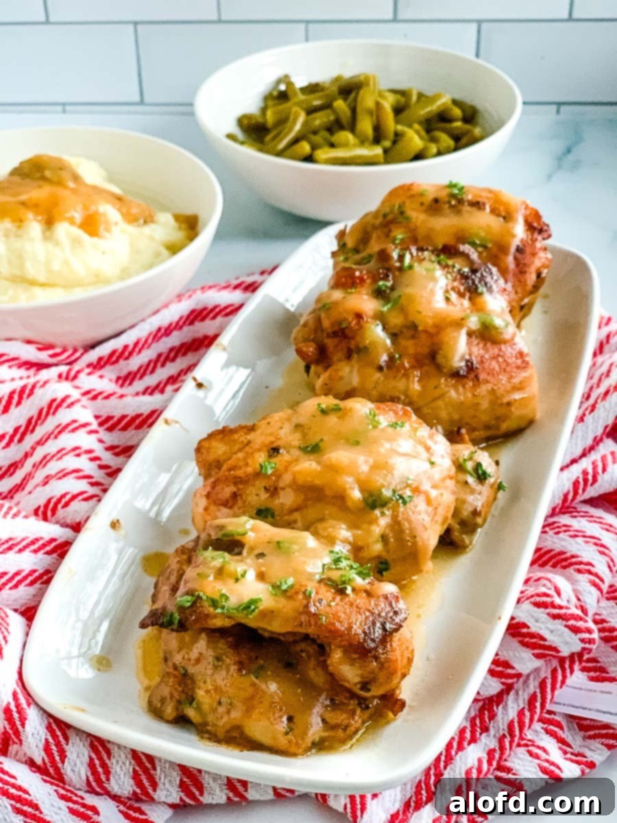 Homestyle Gravy Chicken Thighs 2 A white platter showcasing perfectly pan-seared boneless chicken thighs, golden brown and glistening with gravy, with a side of vibrant green beans and a bowl of creamy mashed potatoes blurred in the background.