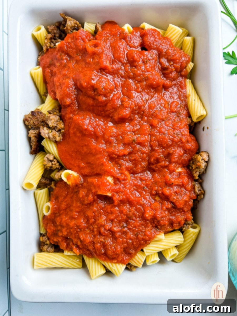 Assembling a sausage rigatoni casserole, showing pasta, sausage, and sauce being mixed.